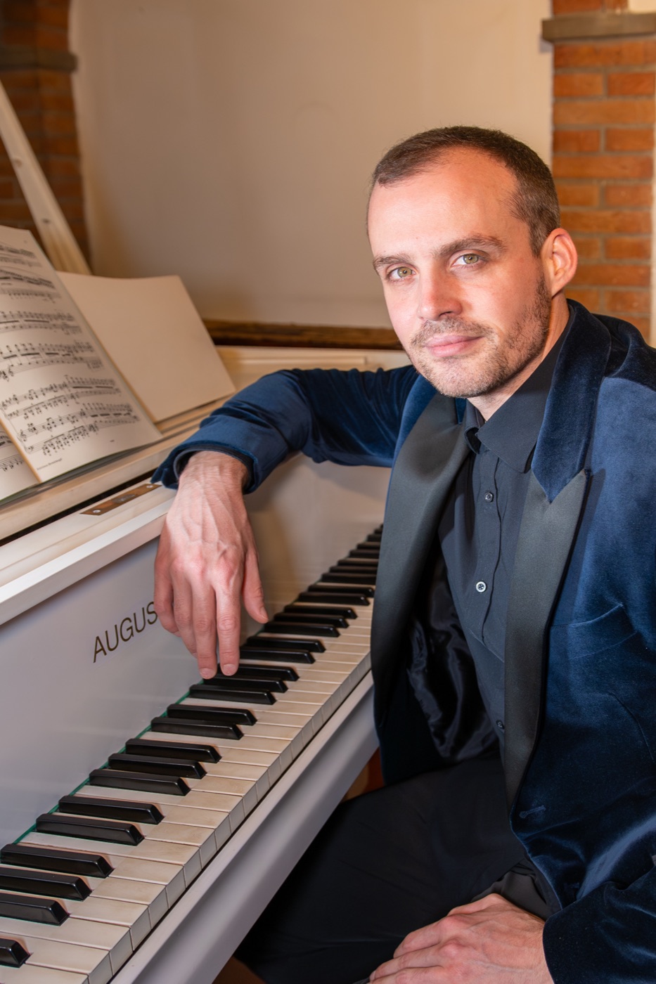 Ludovico Troncanetti seated at a white grand piano with an open score, one hand on the keyboard, photographed by Roberto Bassan